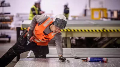 A male in a high vis jacket and hard hat holding a power tool crouches over a pallet.