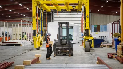 A person wearing a high vis vest and hard hat carries a wooden block through a manufacturing facility passed a forklift.