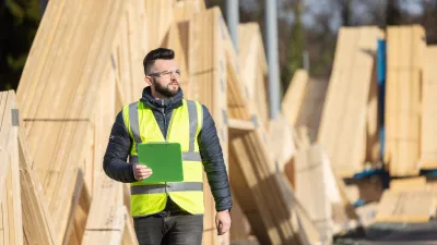 A male with protective goggles and a high-vis jacket carrying a clipboard passed piles of lumber. 