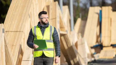 A male with protective goggles and a high-vis jacket carrying a clipboard passed piles of lumber. 