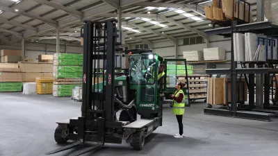 A person in a high vis vest with ear muffs show a tablet screen to a person driving a forklift.