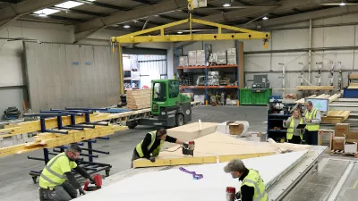 Three people in high vis jackets use power tools to cut wooden frames while two people in high vis jackets with a tablet observe.