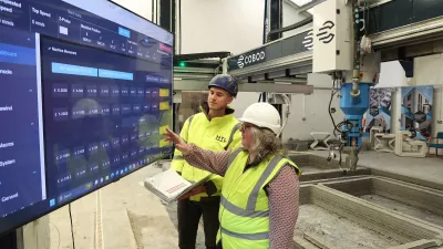 Two people in high-vis vests reviewing data on a large screen, with a 3D printer visible in the background.