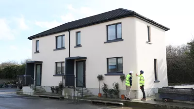 Two people in high vis jackets and hard hats inspect modern white semi-detached houses.