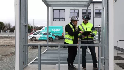 A male and a female read from a tablet inside a prefabricated steel frame set in a car park.