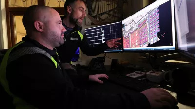 A male points out something in a digital building design on a computer screen to another male.
