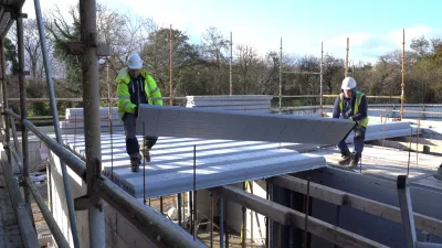 Two people in high vis jackets and hard hats carry a flooring panel to be installed on the second floor of a house.