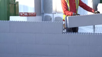 A person in a red jacket and a high vis jacket carries a section of a wall to be clipped to other wall components. 