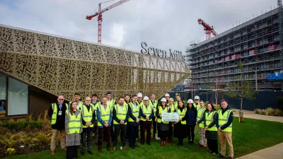 A group of people in high vis vests stand outside a modern structure with a sign reading Seven Mills above and a partially constructed apartment building in the background.