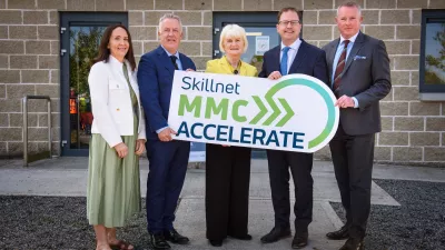 Five people standing outside a stone building, smiling and holding a large sign that reads 'Skillnet MMC Accelerate' with a green and blue logo.
