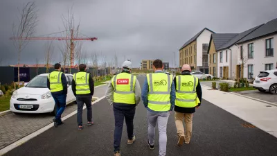 A group of five people in high vis vests walk past a white car through a modern housing development.
