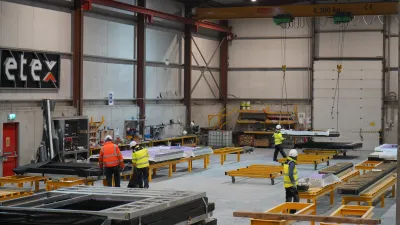 Several people in high vis vests work at machinery stations in a manufacturing facility.