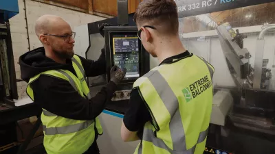 A male with a high vis vest and gloves explains digital readings on a machine screen to another person in a high vis jacket.