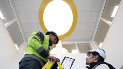 A male in a hard hat with a tablet shows the screen to another male in a hard hat standing on a ladder. 