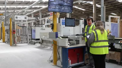 Two workers in high-vis vests monitoring a 3D printer control station inside a large manufacturing facility.