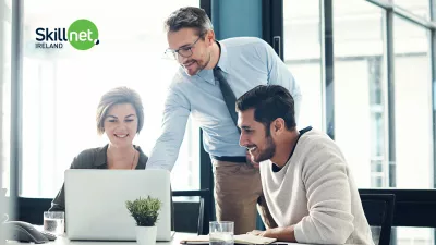 Three colleagues working together in a bright modern office, gathered around a laptop and discussing something on the screen.