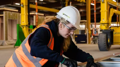 Person in high-vis vest and hard hat inspecting the quality of a metal frame in a factory.