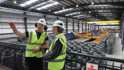 A male with a high vis vest, hard hat, and ear muffs points at something to a female in a high vis vest, hard hat, and ear muffs.