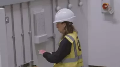 Electrician in high-vis vest and hard hat working on equipment near a control panel.