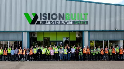 A large group of people stands outside a corrugated metal building with a Vision Built sign over the entrance.