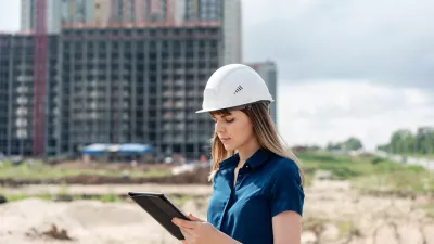A female in a hard hat with a blue top reads a tablet on a construction site.