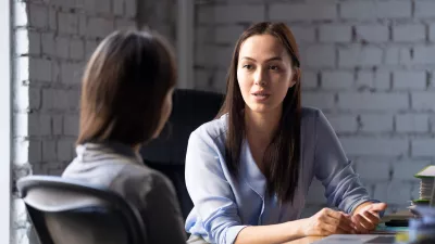 Two females in business clothing sit at a desk in a modern office space talking.