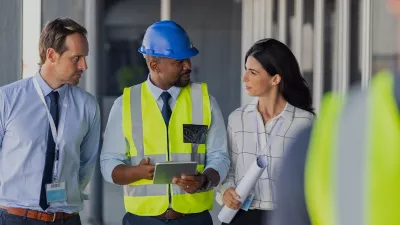 A male in a hard hat and high vis jacket holding a tablet explains something to two people in business clothing.