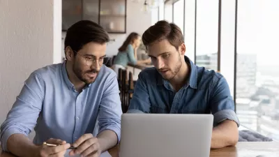Two males in business shirts sit at a desk and read from a laptop. One takes notes with a pen on a piece of paper.