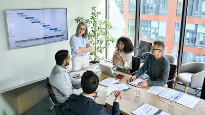 A group of people in business clothing with laptops and pieces of paper in front of them sit around a long table, talking.