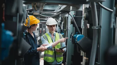 Two people in hard hats and work overalls, one holding a clipboard and the other holding a tablet, inspect machinery.