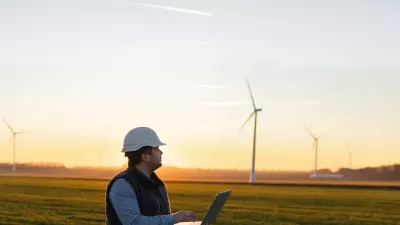 A male in hard hat and vest with an open laptop stands in a field with wind turbines in the background.