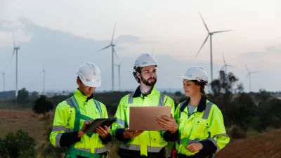 A group of people in high vis jackets and hard hats read from tablets standing in a field with wind turbines.