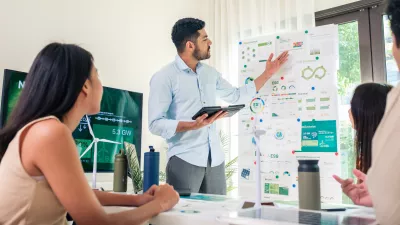 A male in a business shirt stands with a tablet in his hand explaining information on a flip chart to a group of people sat at a table. 