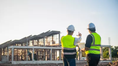 Two people in high-vis vests and hard hats standing at a construction site, looking at a building under development.