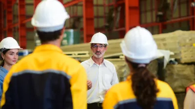 A male with glasses and a hard hat addresses a crowd of people wearing work overalls and hard hats.