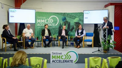 A panel of five speakers seated on a stage during a conference, with one person standing and speaking. Two screens and a green BIM Gazelle backdrop are visible behind them.