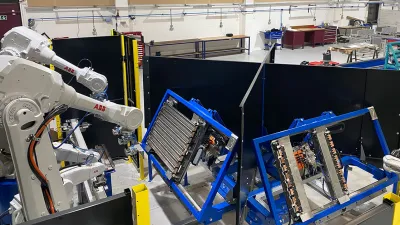Industrial robotic arms working on components mounted in blue frames inside a manufacturing or training facility. Workbenches and tools are visible in the background.