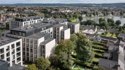 Aerial view of a modern apartment complex with multiple grey buildings, surrounded by traditional houses and greenery, with a lake and town visible in the background.