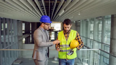 Two people wearing hard hats are standing on a construction site, discussing a project.
