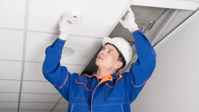Worker in hard hat installing or inspecting a ceiling smoke detector.
