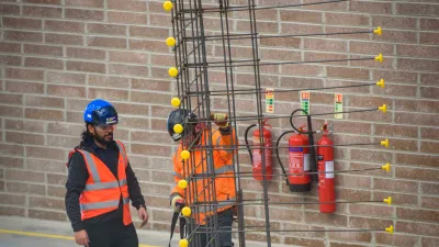 Two construction workers in safety gear standing near a vertical rebar structure inside a building under construction.