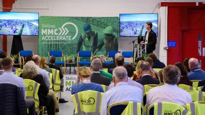 Audience at an indoor event, watching a panel discussion on a stage with presentation screens and green construction-themed visuals in the background.