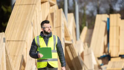 Construction worker wearing a high-visibility vest and holding a clipboard, standing in front of wooden roof trusses at an outdoor building site.