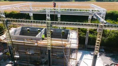 Aerial view of a construction site where a large 3D concrete printer is building two small rectangular structures.