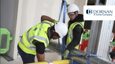 Two construction workers in high vis jackets, white hard hats, and goggles work on tying a pipe
