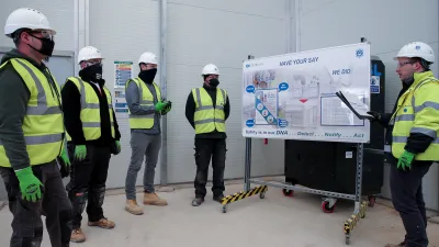 Four construction workers in hard hats, high vis jackets, and face masks listen to another worker doing a safety presentation from a whiteboard