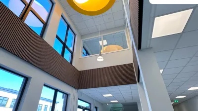 A bright, modern school interior with large windows, a central skylight featuring a yellow circular frame, and a staircase leading to the upper floor.