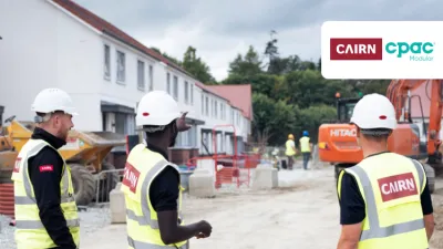 Three construction professionals in safety gear and hard hats are standing on a residential building site, discussing progress. Houses under construction are visible in the background.