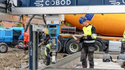 A construction worker in a hard hat and high-vis jacket observes a 3D printer building a concrete wall