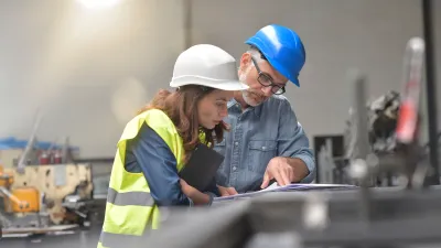 Two people wearing hard hats and safety vests reviewing a technical drawing in an industrial setting.